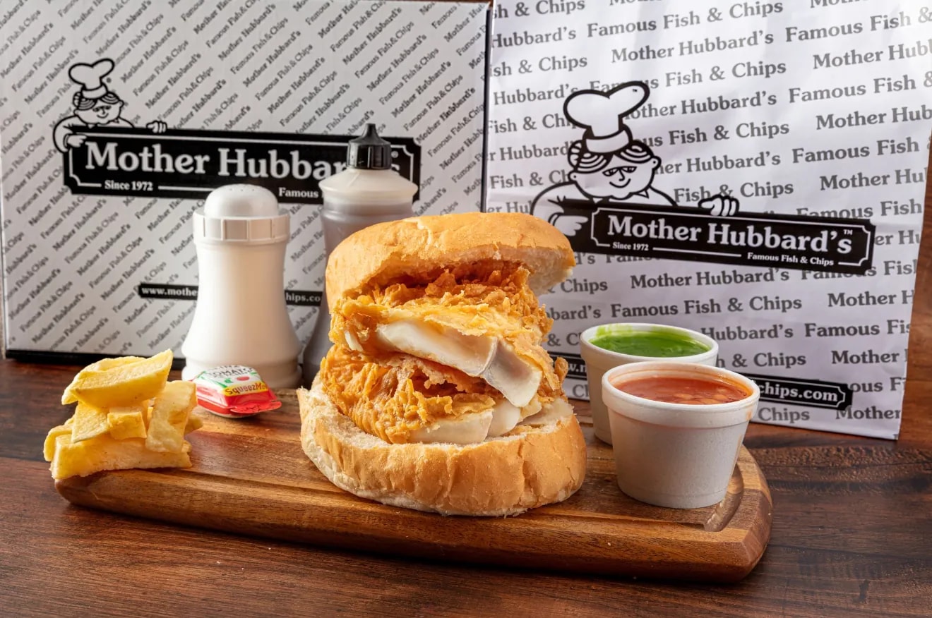 A battered fish butty in a soft roll on a board, with lemon, chips and two dips