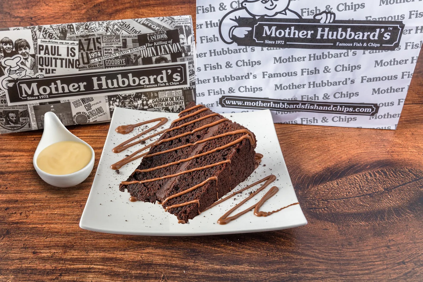 A slice of chocolate cake drizzled with chocolate sauce on a square plate, with a pot of custard
